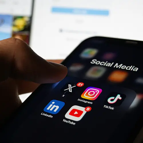 Smartphone screen showing social media apps including Instagram, TikTok, YouTube, LinkedIn, and X, with notification badges visible. A finger hovers over the screen, symbolizing active engagement. Represents the digital tools beginner fitness coaches use to get their first clients, optimize content, and book sales calls through platforms like Instagram and DMs.