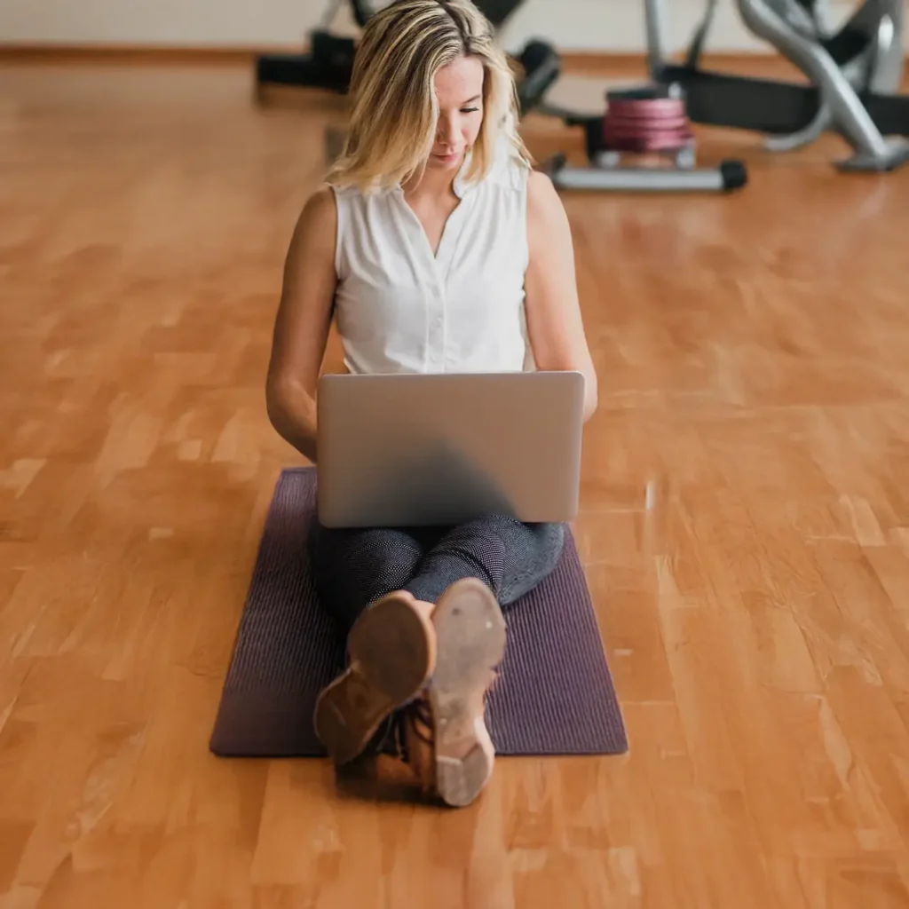 Female coach with a white blouse working on her laptop on a yoga mat on a wooden floor in a gym.