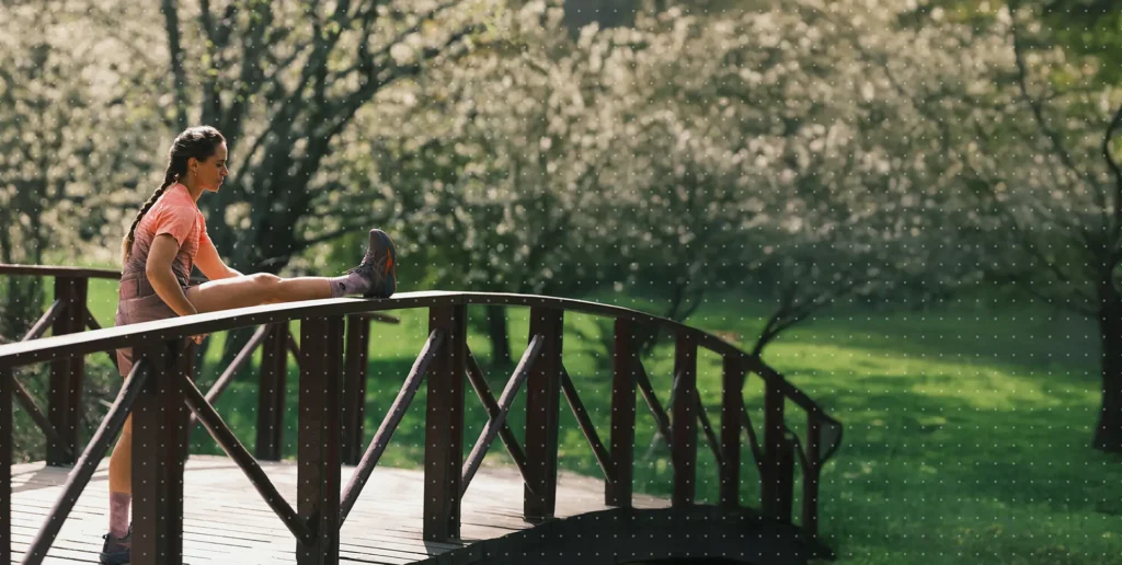 Anna Haag stretching on a bridge in sportswear, with blurred trees in bloom and grass in the background.